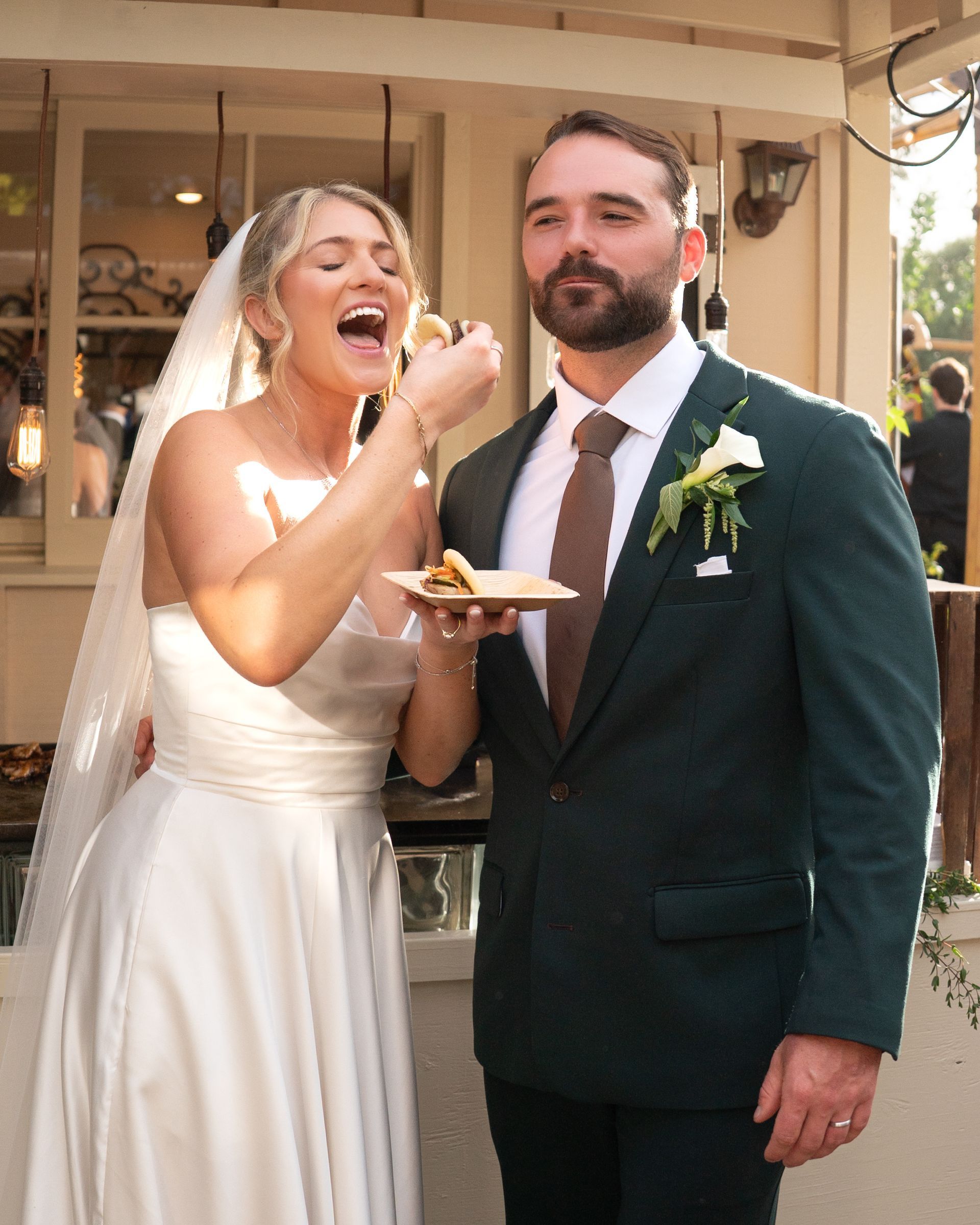 Bride feeding cake to groom at a wedding reception outdoors, both in formal attire.