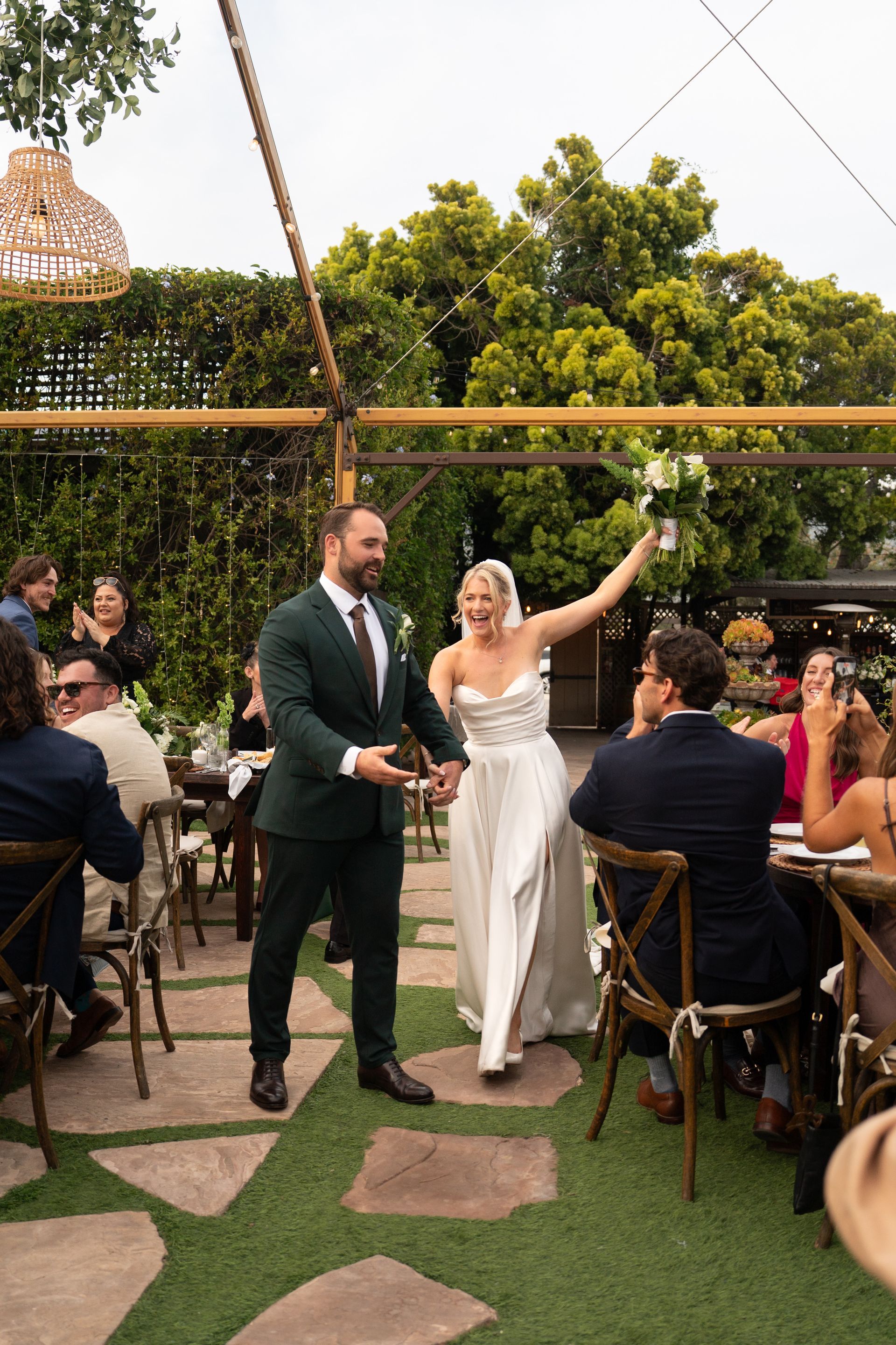 Bride in white waving at an outdoor wedding reception beside the groom in a dark suit