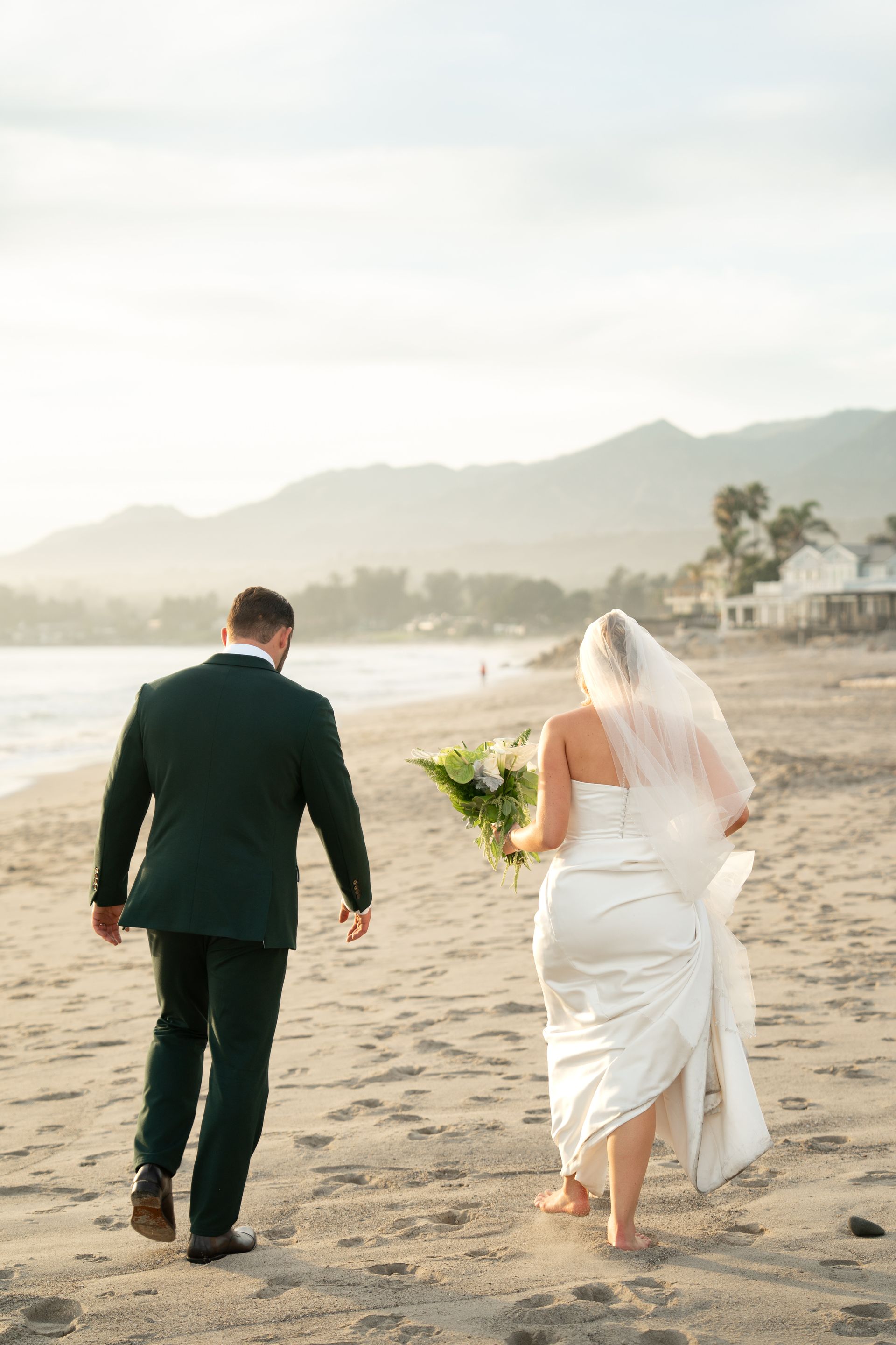 Couple walking barefoot on a sandy beach, bride in white dress and veil holding flowers beside groom in dark suit