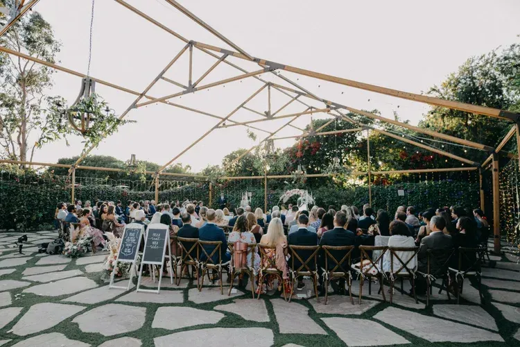 Outdoor wedding reception under a geometric canopy with seated guests around a long banquet table.