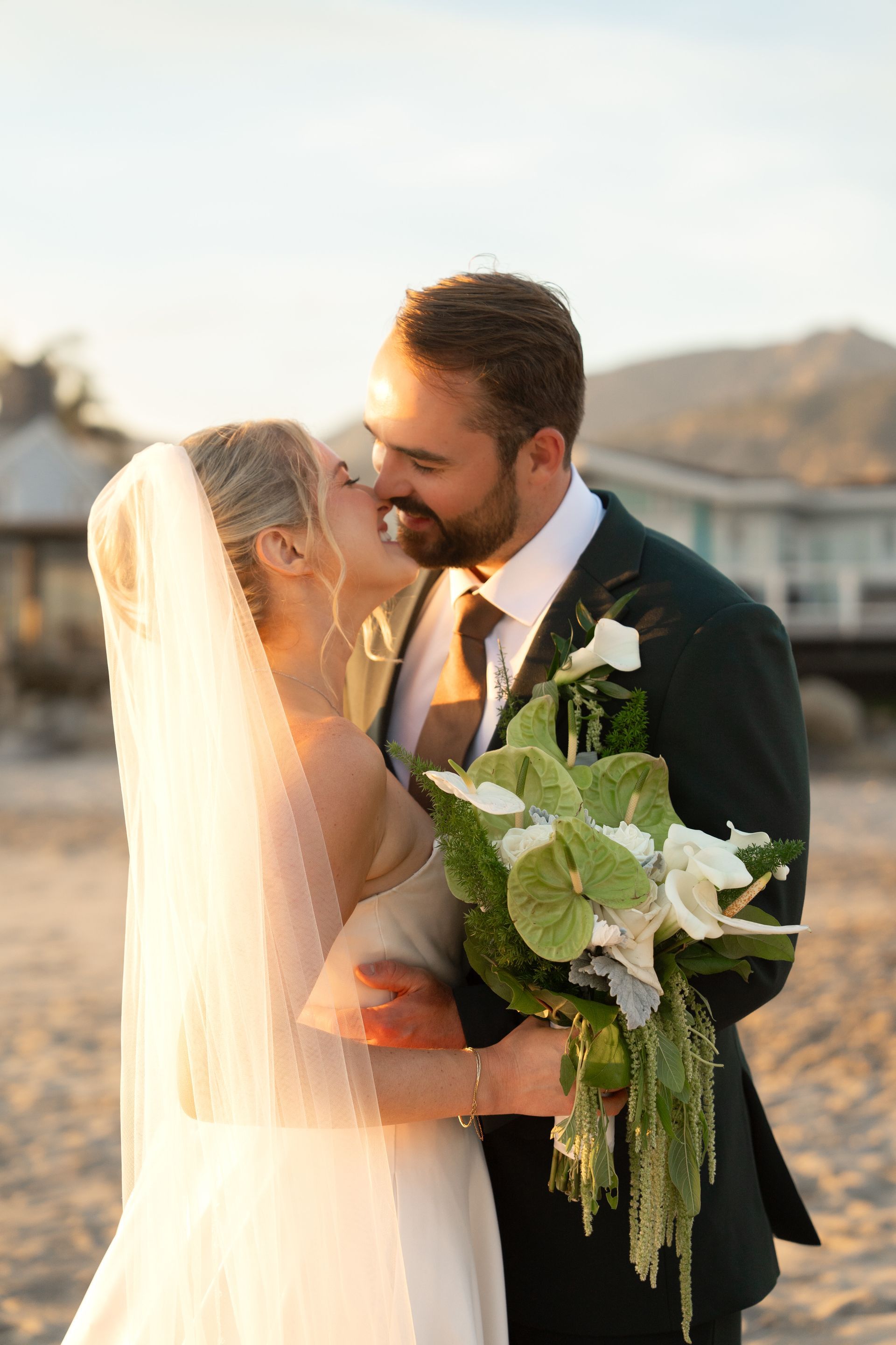 Bride and groom embrace outdoors at sunset, with the bride holding a bouquet of greenery.