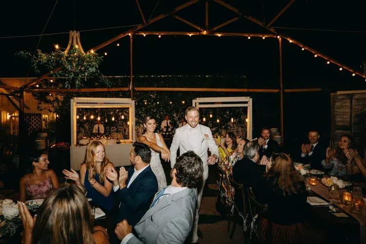 Outdoor evening wedding reception with guests dining at candlelit tables and applauding a couple under string lights