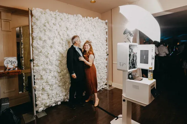 Couple posing at a photo booth beside a white floral wall, with a large studio light in front.