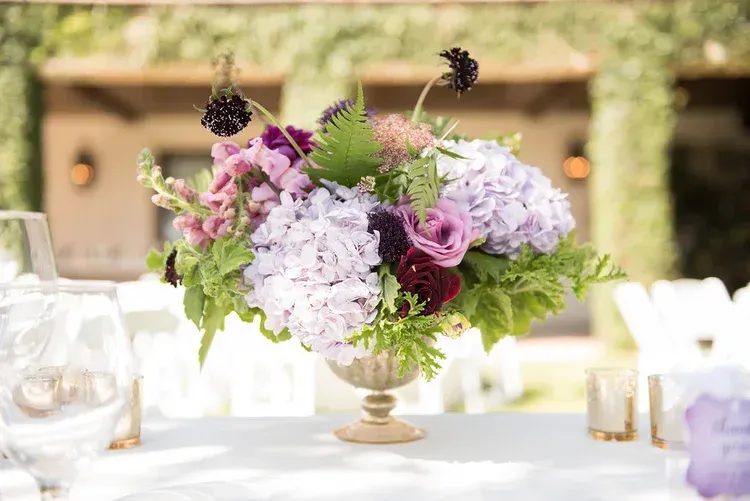 Lavender and purple floral centerpiece in a gold vase on a white table at an outdoor event