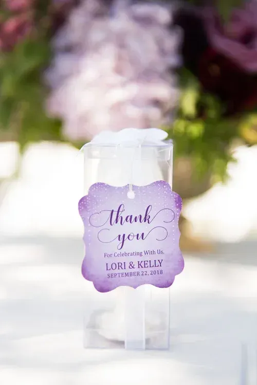 Purple “Thank you” wedding favor tag on a white bag, with blurred flowers in the background