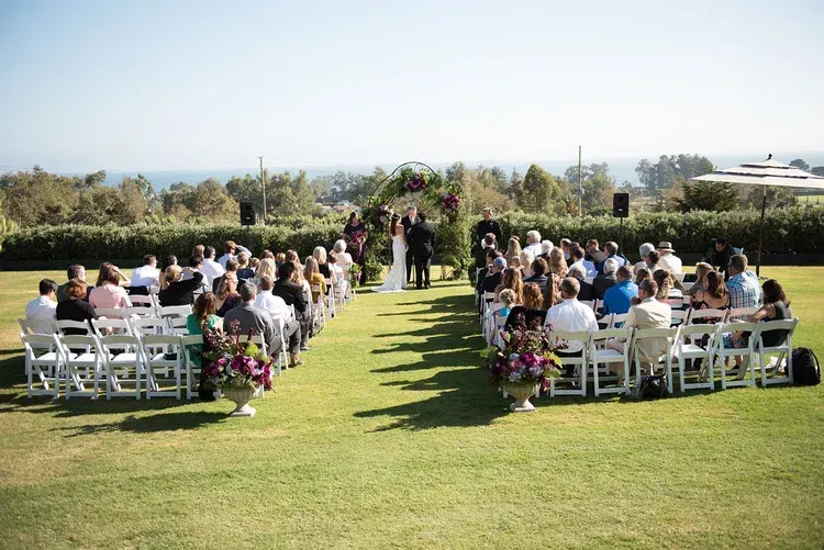 Outdoor wedding ceremony with guests seated in white chairs on a grassy lawn aisle