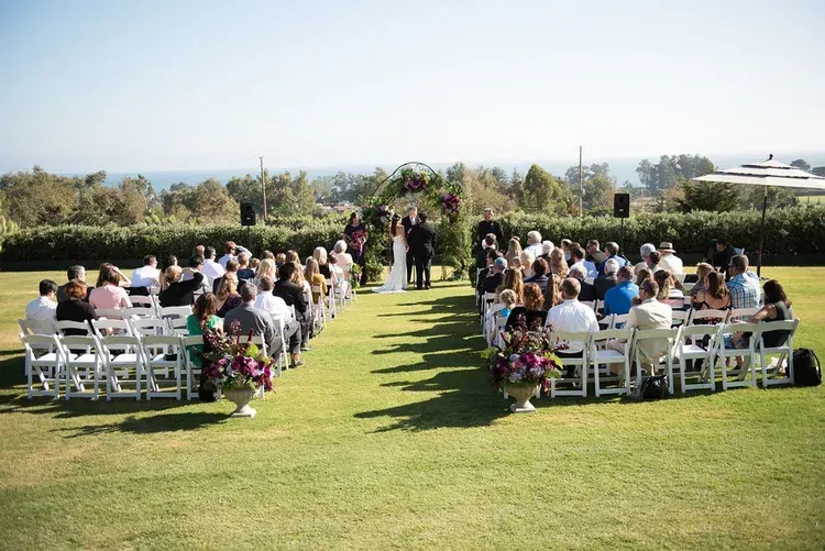 Outdoor wedding ceremony with guests seated on white chairs along a grassy aisle