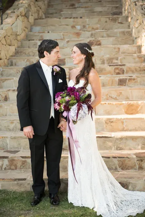 Bride and groom stand on stone steps, smiling and holding a purple bouquet.