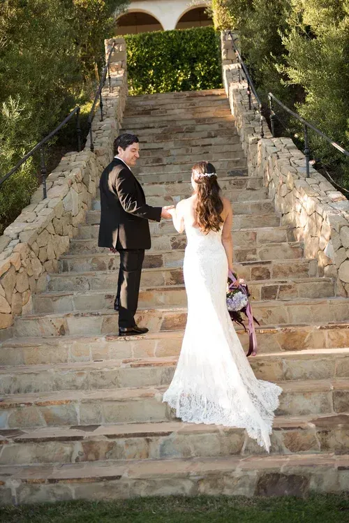 Bride and groom holding hands on stone staircase outdoors, bride in white gown with bouquet, groom in dark suit
