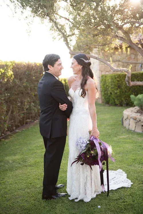 Bride and groom in wedding attire standing together outdoors, holding a bouquet and facing each other.