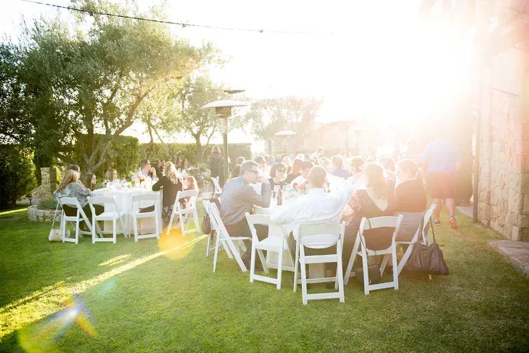 Outdoor wedding reception with guests seated at white tables under trees in bright sunlight.