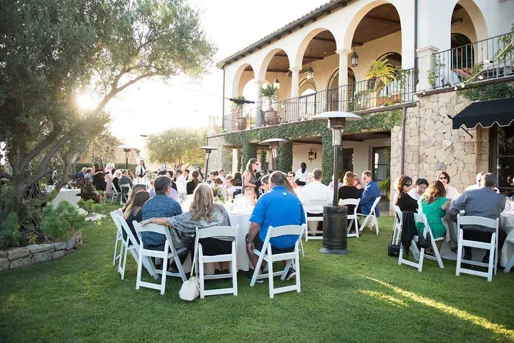 Outdoor gathering on a grassy lawn with guests seated at tables beside an arched stone building