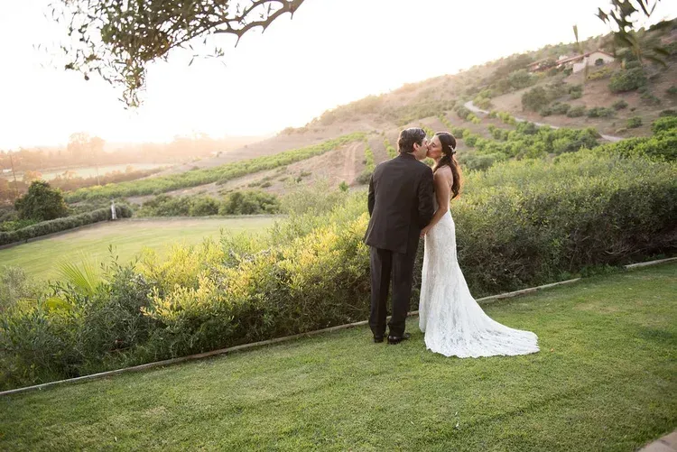 Bride and groom embracing on a grassy hillside at sunset, with a long white train and lush valley behind them