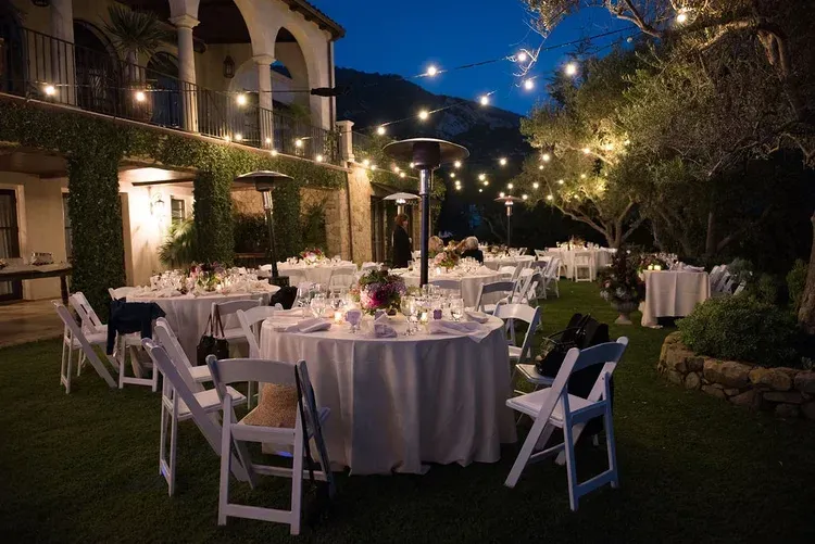 Elegant outdoor dinner setup with round tables, white chairs, and string lights in a garden courtyard at dusk