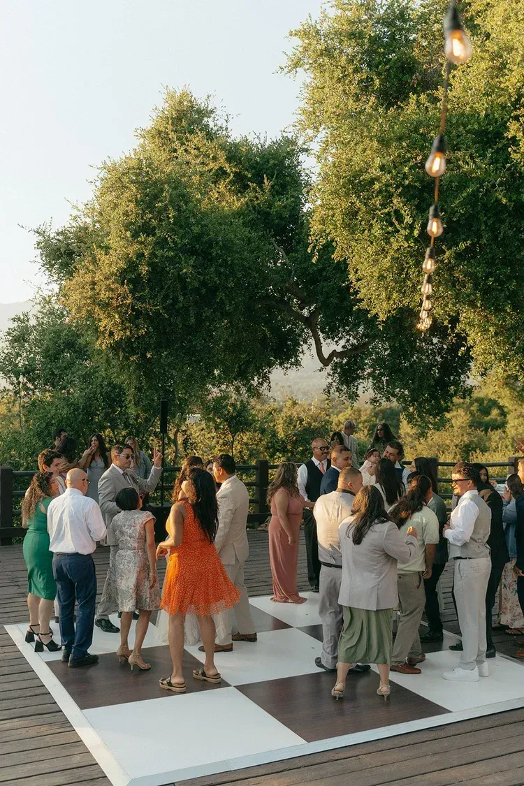 Guests mingling at an outdoor wedding reception on a white dance floor under string lights and trees