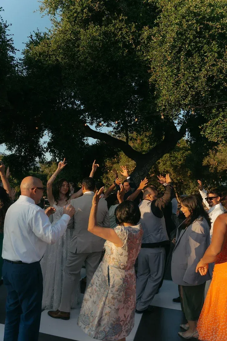 Guests dancing and raising hands at an outdoor celebration under a large tree
