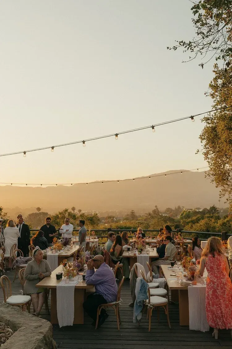 Outdoor sunset dinner gathering with long communal tables, string lights, and people seated among vineyards.