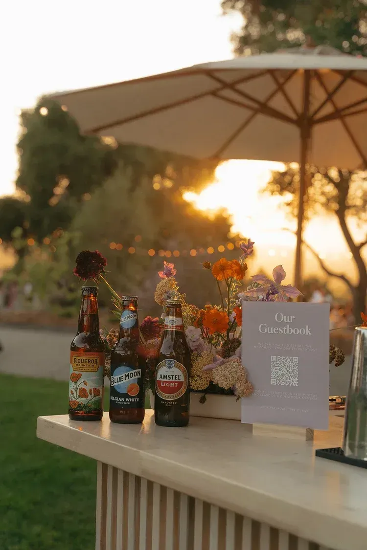 Decorated outdoor table with beer bottles, flowers, and a sign at sunset