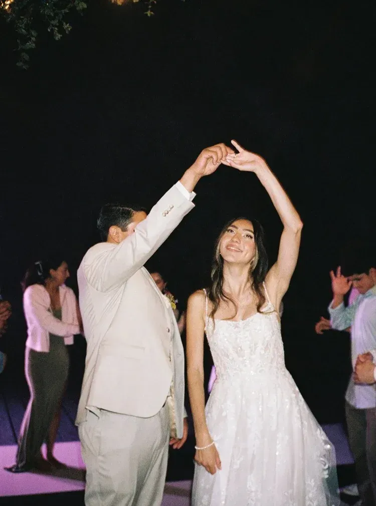 Couple dancing under an arched hand at an outdoor evening celebration, with guests in the background