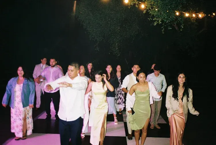 Group walking and dancing outdoors at night under string lights, with one person leading the way.