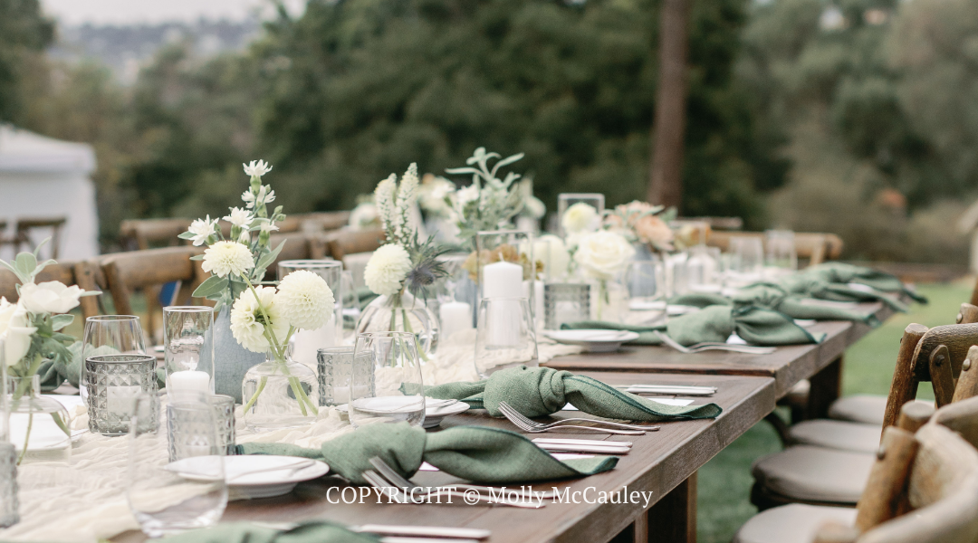Elegant outdoor dining table set with white flowers, candles, and green napkins in a garden setting