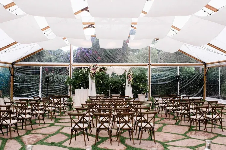 Open-air event tent with round tables and wooden chairs under white draped fabric and hanging greenery