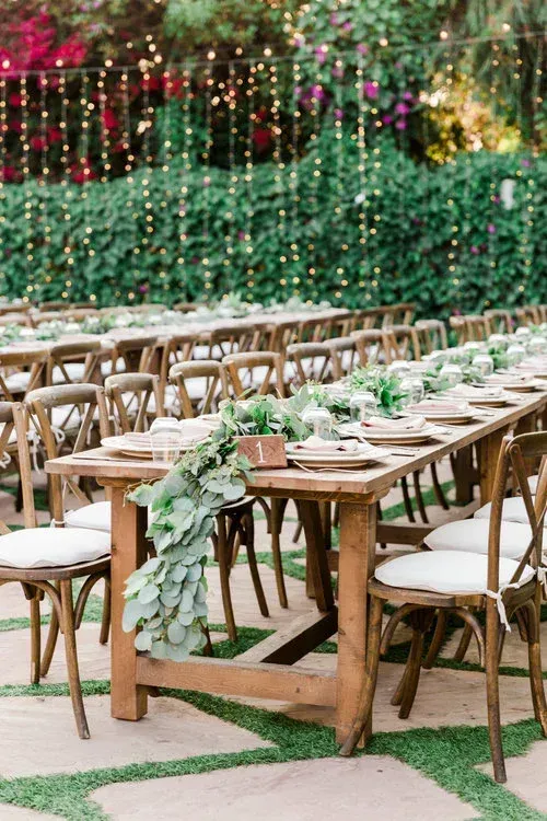 Outdoor wedding reception setup with wooden tables, white chairs, and green floral garlands