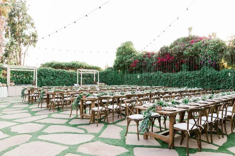 Outdoor patio with wooden tables and chairs on stone paving, surrounded by green hedges and flowers