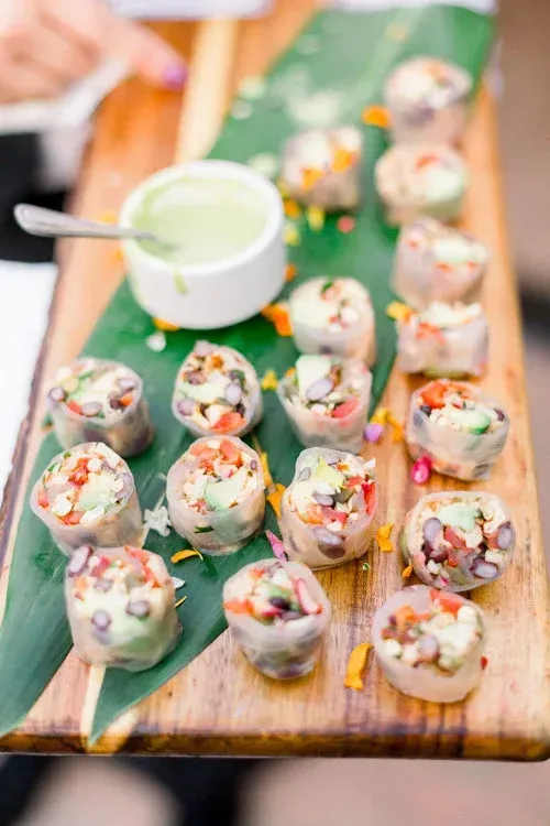 Wooden board with rows of vegetable sushi rolls and a small bowl of green dipping sauce