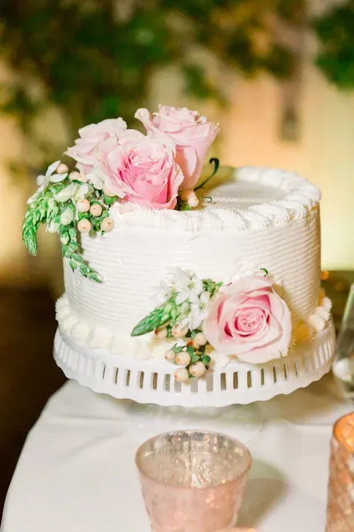White frosted cake with pink roses and greenery on a table, decorated for a celebration