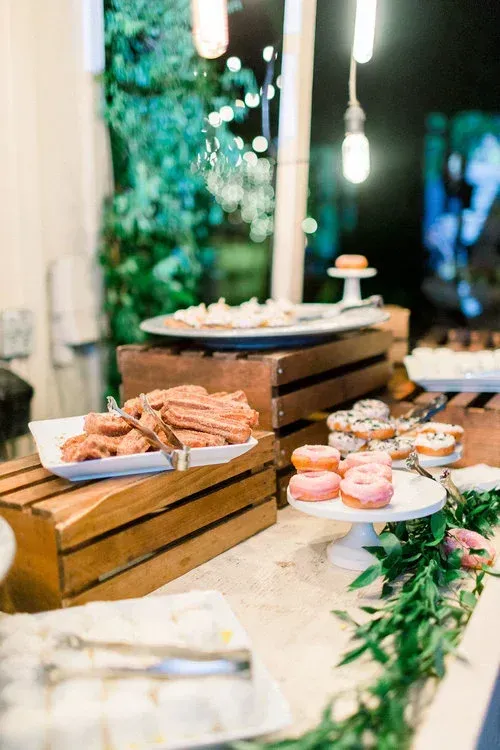 Dessert buffet with pastries and cookies on wooden crates, decorated with greenery at a softly lit event.