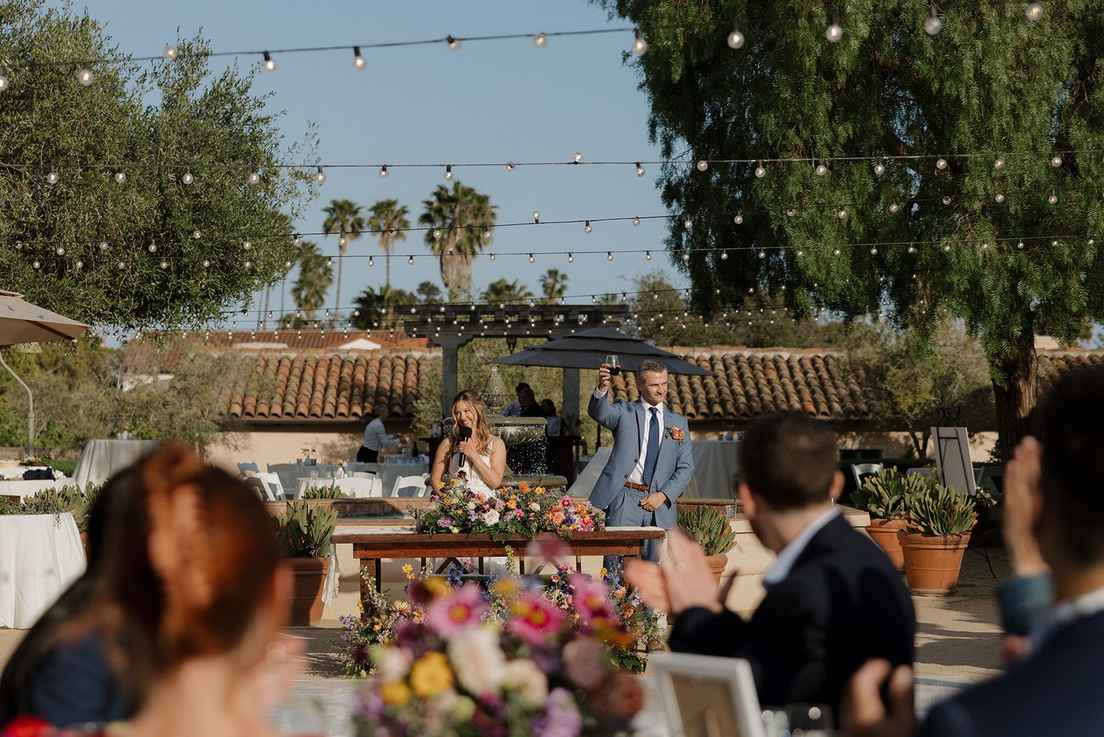 Outdoor wedding ceremony with couple at altar, guests seated, and floral decorations under string lights.