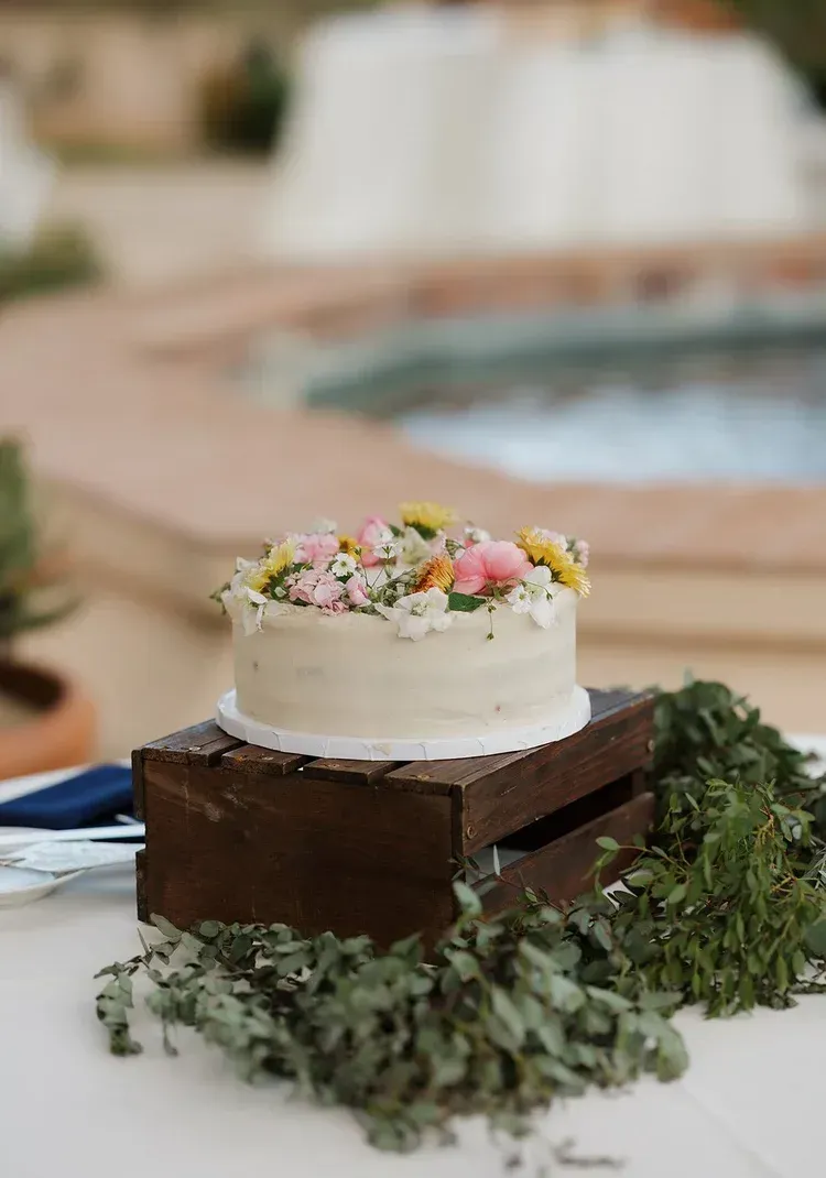 Small white frosted cake with pink flowers on a wooden box, set on a table outdoors near a pool.