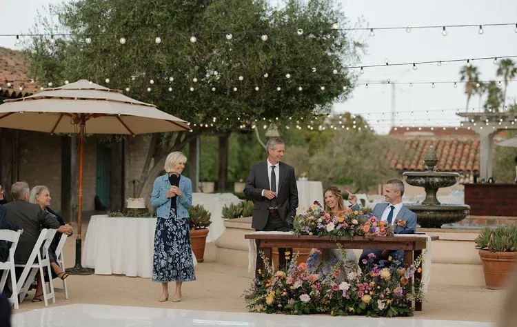 Outdoor wedding ceremony with speaker at flower-decorated table and guests under string lights