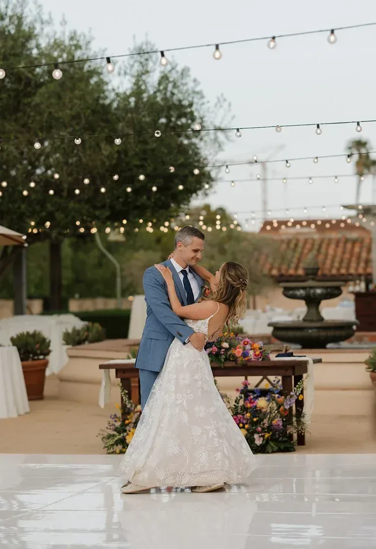 Couple dancing at an outdoor wedding reception under string lights, with floral decor and fountain in the background