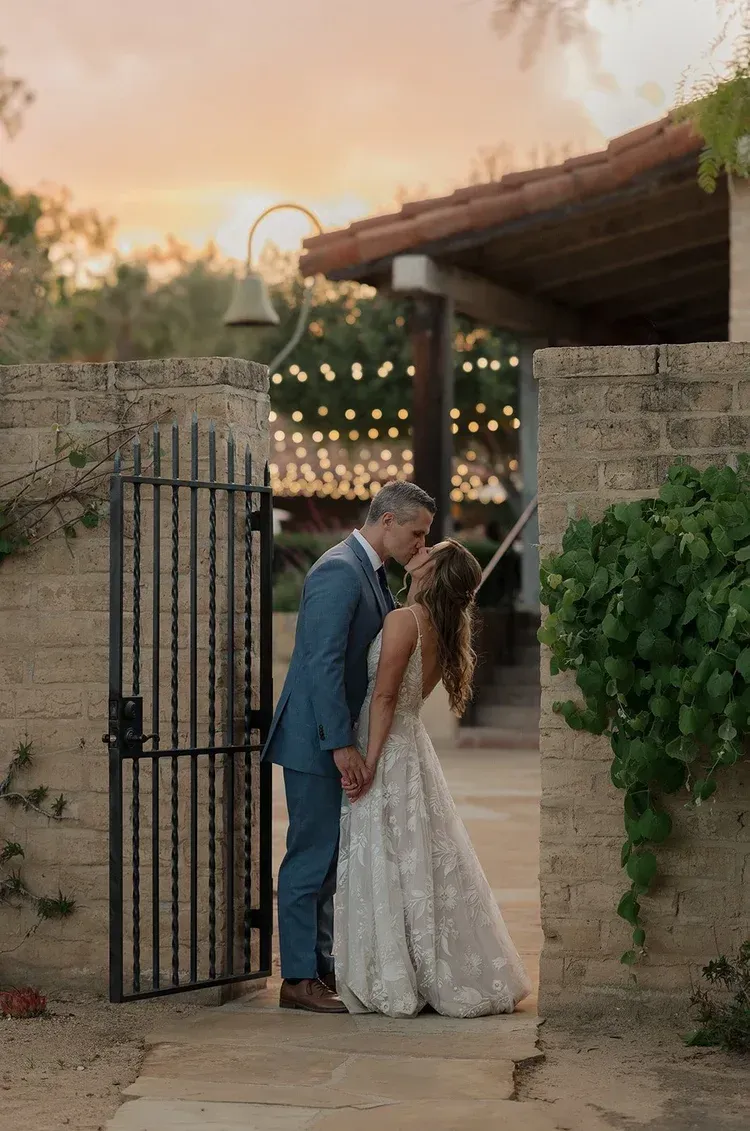 Newlyweds kissing at a garden gate at sunset, bride in a white lace gown, groom in a blue suit