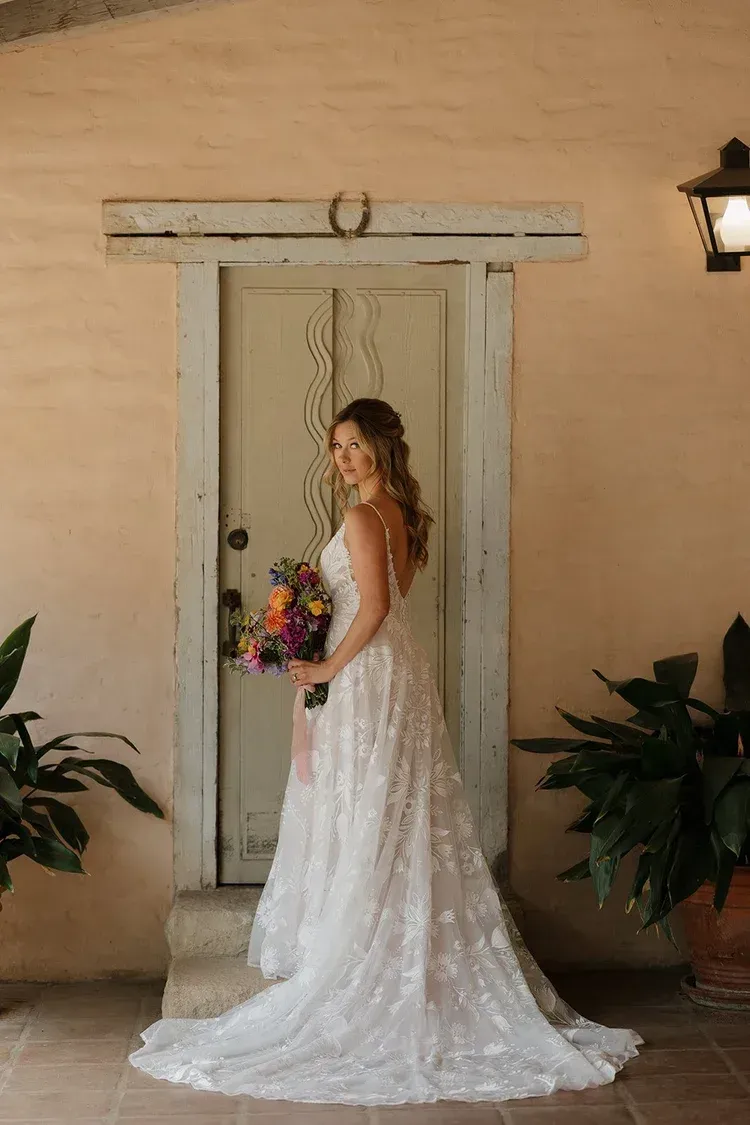 Woman in a white lace wedding dress holding a bouquet, standing before a rustic doorway.