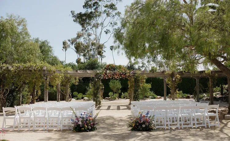 Outdoor wedding ceremony setup with white chairs, floral arrangements, and a vine-covered pergola under trees