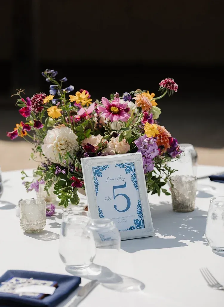 Table centerpiece with colorful flowers and a blue-framed number 5 card on a white tablecloth.