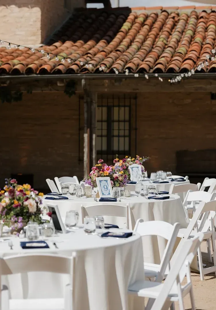 Outdoor patio set with round white-draped tables and white chairs, decorated with colorful floral centerpieces.