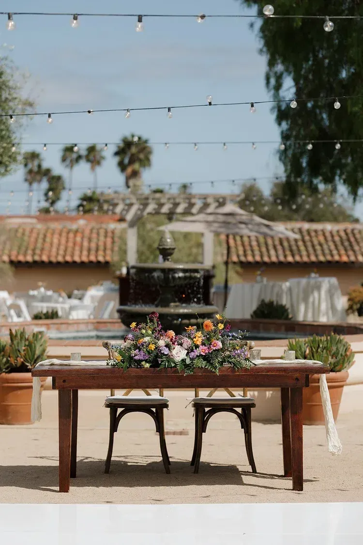 Outdoor dining table with flower centerpiece and two chairs in a sunny courtyard