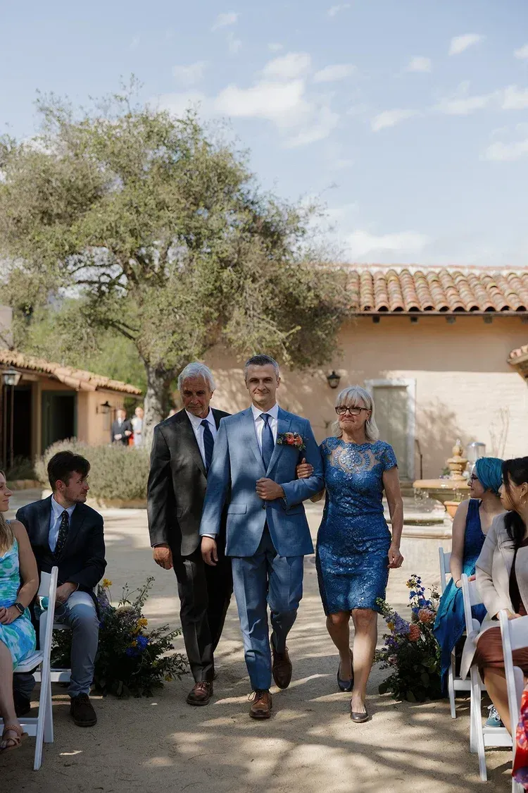Three people in blue formal wear walking down an outdoor aisle at a wedding ceremony.