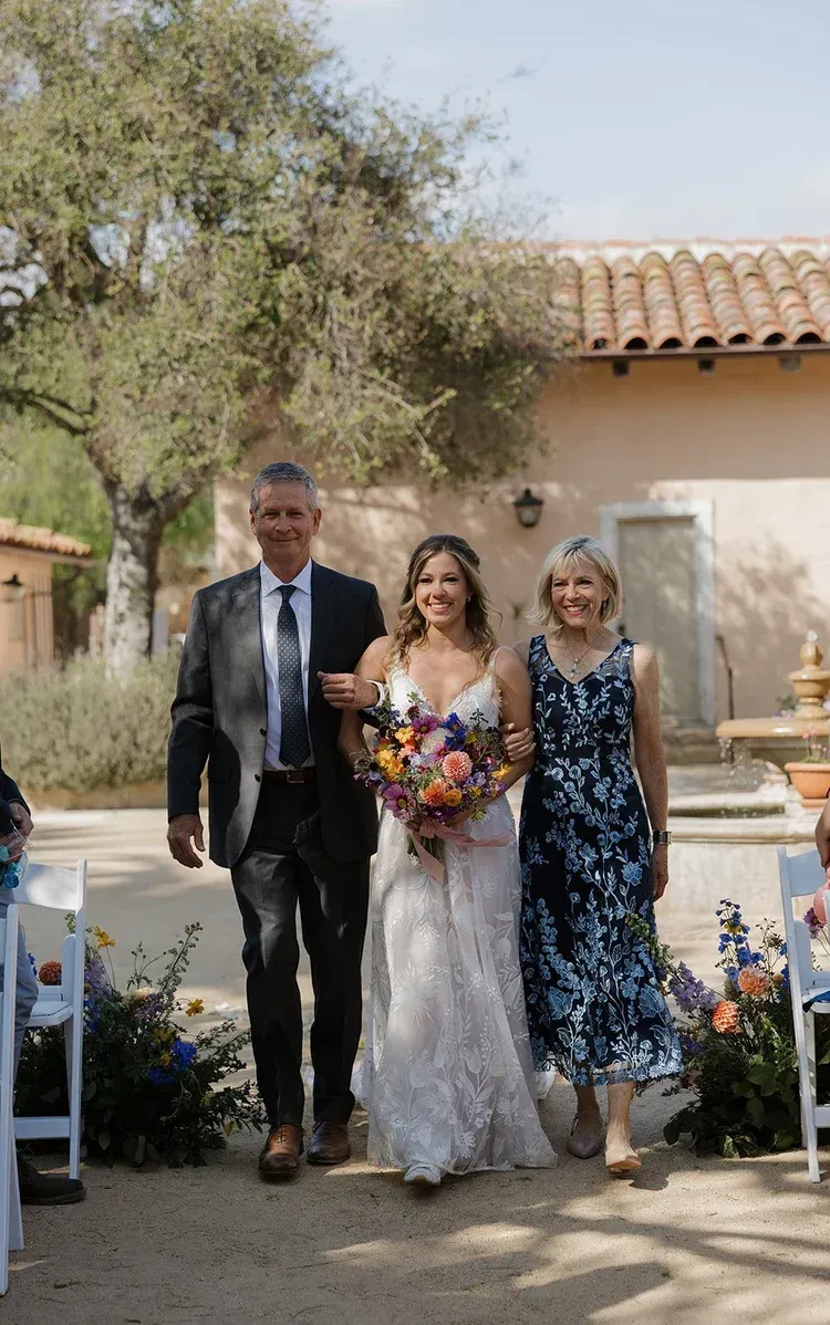 Bride in white gown walks with two companions, holding bouquet, in an outdoor courtyard wedding ceremony.