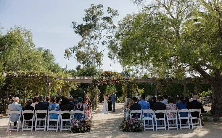 Outdoor wedding ceremony with seated guests under white chairs, surrounded by greenery and trees
