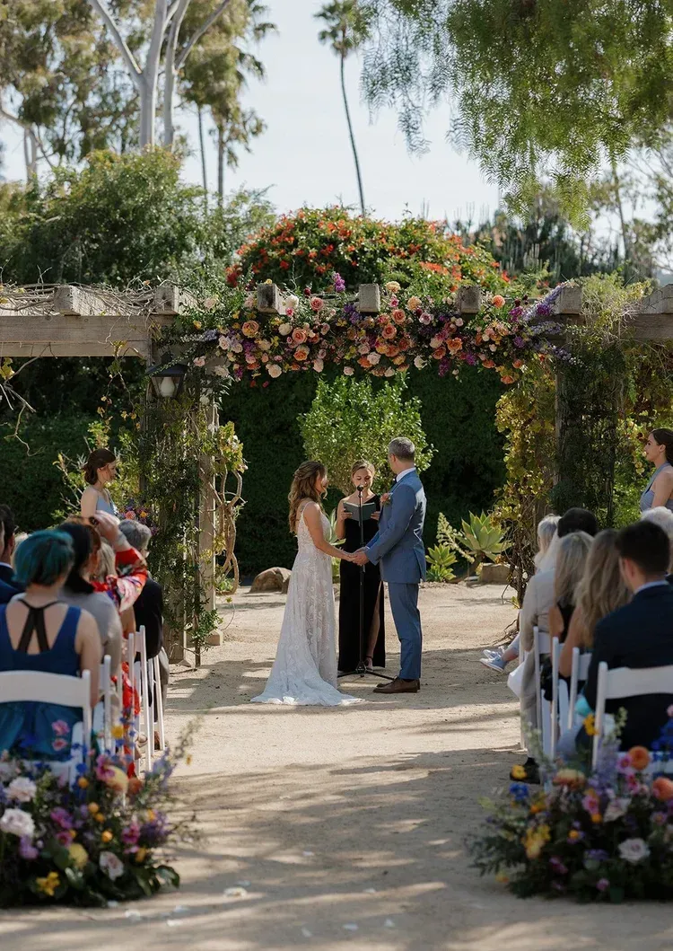 Outdoor wedding ceremony under a flower-covered arch, with couple exchanging vows and guests seated along the aisle