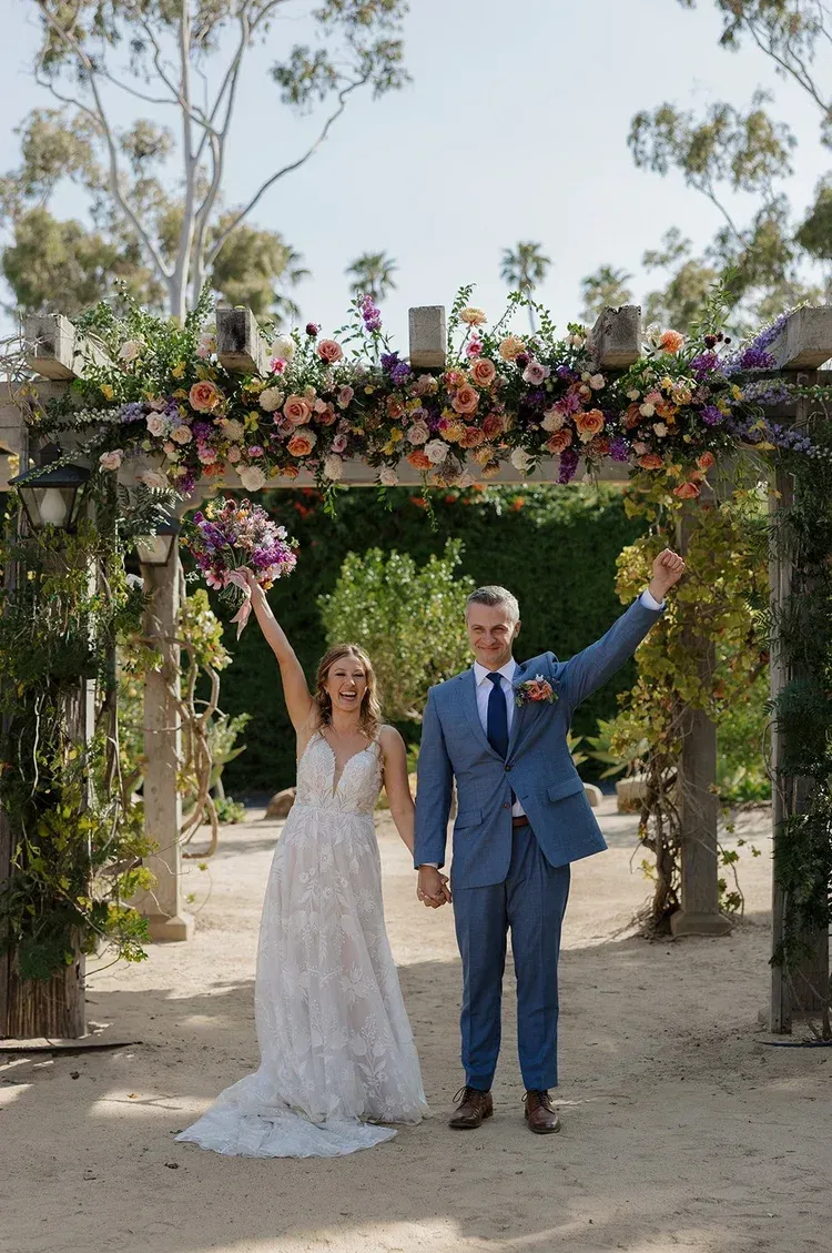 Newlyweds under a flower-covered arch, smiling and raising their arms outdoors.