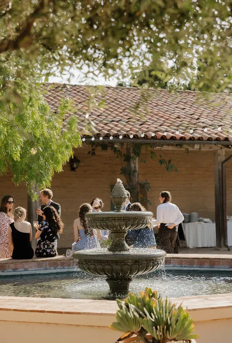 Courtyard fountain with people standing nearby under trees beside a tiled-roof building
