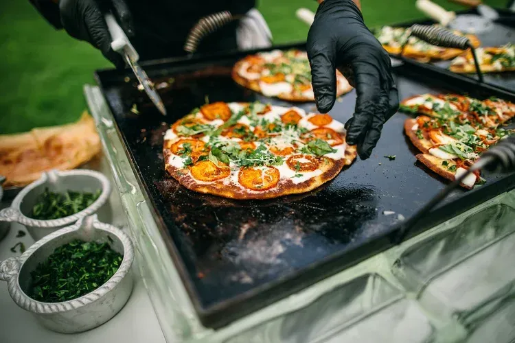 Gloved hands place a vegetable pizza on a black griddle at an outdoor cooking station