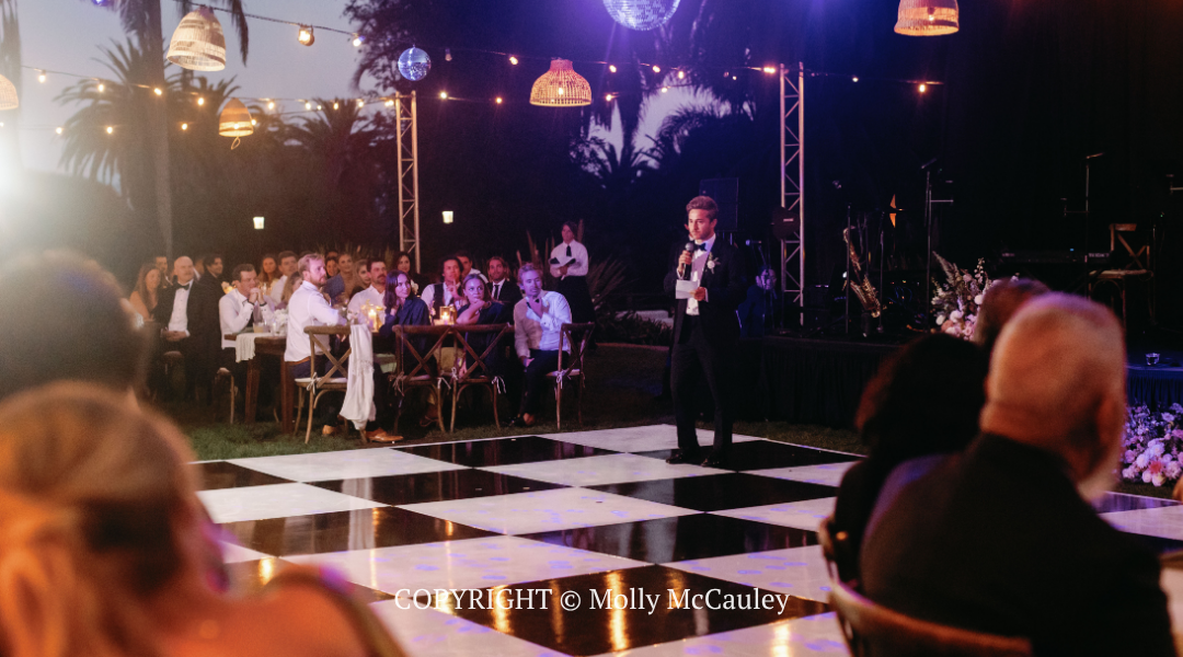 Speaker addressing seated audience on a black-and-white dance floor at an evening event