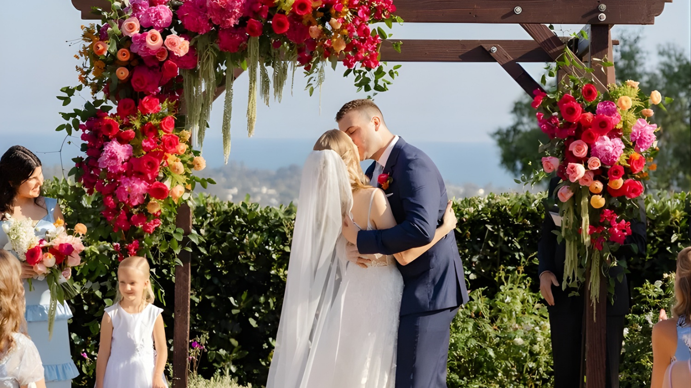 Bride and groom kissing under a flower-covered arch at an outdoor wedding ceremony